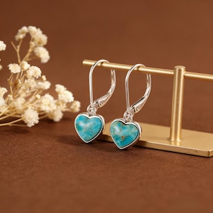 May include: Silver heart-shaped earrings with turquoise centres. The earrings have a lever-back closure and are displayed on a gold stand. The background is a warm brown.
