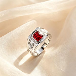 May include: A silver ring with a square, deep red gemstone at the center. The ring is accented with small, clear stones and has a polished finish. The ring is set against a soft, cream-colored background.