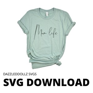 May include: A light green t-shirt with the words "Mom life" in a black script font. The shirt is knotted at the bottom and has rolled-up sleeves. The text "DAZZLEDDOLLZ SVGS" and "SVG DOWNLOAD" are at the bottom of the image.