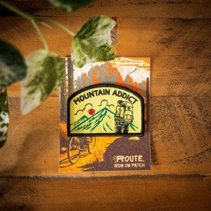 May include: A yellow and green iron-on patch featuring a hiker with a backpack in front of a mountain range. The patch reads "Mountain Addict" and "Route. Iron On Patch".
