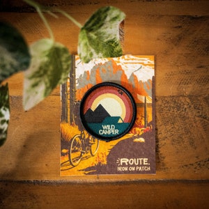 May include: A black and green embroidered iron-on patch with a sunset and mountains design. The patch says "Wild Camper" and "ROUTE. IRON ON PATCH".