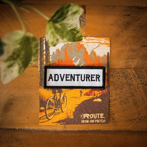 May include: A white iron-on patch with black text that reads "Adventurer" on a brown background with a mountain scene and a bicycle.