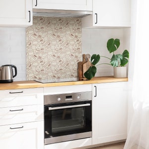 Puede incluir: Una cocina blanca con un horno de acero inoxidable y un protector contra salpicaduras con un patrón marrón y blanco de varios panes y pasteles.