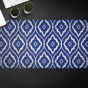 Op de afbeelding: Een rechthoekige bureaumat met een blauw en wit geometrisch patroon. Het ontwerp heeft een herhalend motief van gestileerde, onderling verbonden vormen. De mat ligt op een zwart oppervlak, met een laptop en kleine potplanten zichtbaar.