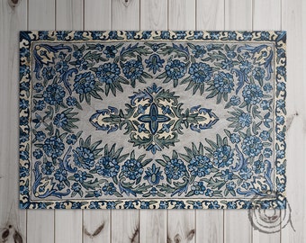 Könnte beinhalten: Rechteckiger Teppich mit floralem Design. Der Teppich hat einen hellgrauen Hintergrund mit einem Rand und einem zentralen Design aus blauen Blumen und grünen Blättern. Der Teppich liegt auf einer hellen Holzoberfläche.