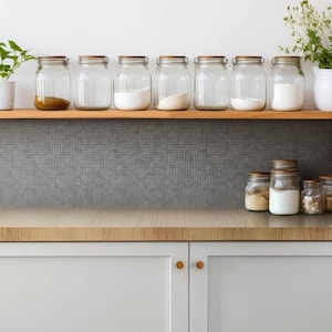 May include: A collection of clear glass storage jars with wooden lids arranged on a wooden shelf and a countertop. The jars contain various white powders, grains, and brown ingredients. The kitchen features light gray cabinets and a gray tiled backsplash.