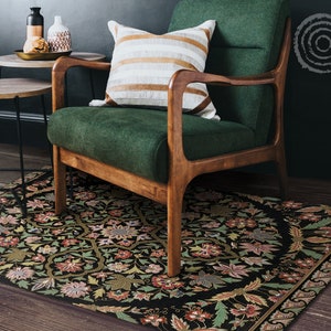 Puede incluir: Un sillón de terciopelo verde con patas y brazos de madera se encuentra sobre una alfombra negra con estampado floral. El sillón tiene un cojín a rayas blancas y marrones en el asiento.