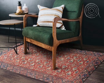 May include: A green armchair with wooden legs and arms sits on a red and cream patterned rug. A small table with a wooden top and black metal legs sits in front of the chair. The chair has a white and brown striped pillow on it.