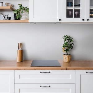 May include: A kitchen scene featuring white cabinets with black handles, a wooden countertop, a black induction cooktop, a wooden knife block with three knives, and a small green plant in a woven basket, set against a light gray tiled wall.