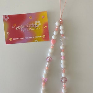Peut inclure: Un porte-cl&eacute;s de t&eacute;l&eacute;phone en perles blanches et roses avec un charme en forme de c&oelig;ur rose et une ficelle rose et blanche. Le charme est attach&eacute; &agrave; un &eacute;tui de t&eacute;l&eacute;phone avec une boucle.