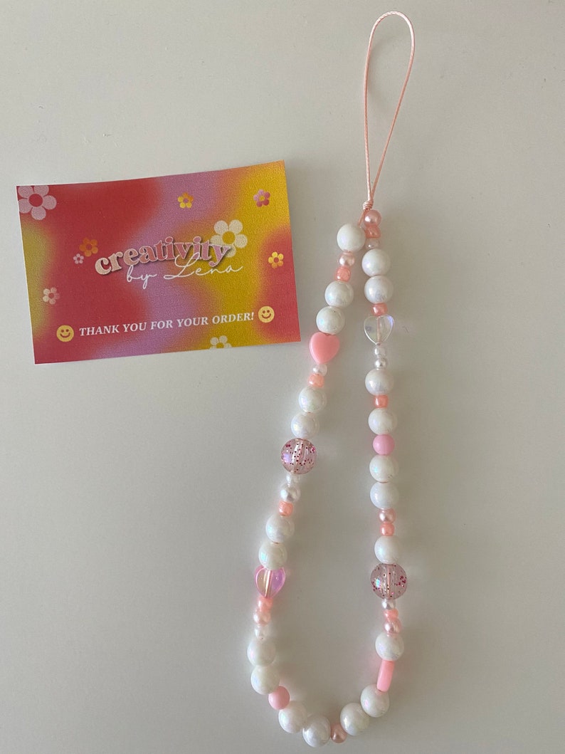 Peut inclure: Un porte-cl&eacute;s de t&eacute;l&eacute;phone en perles blanches et roses avec un charme en forme de c&oelig;ur rose et une ficelle rose et blanche. Le charme est attach&eacute; &agrave; un &eacute;tui de t&eacute;l&eacute;phone avec une boucle.