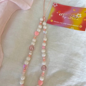 Peut inclure: Un porte-cl&eacute;s de t&eacute;l&eacute;phone en perles blanches et roses avec un charme en forme de c&oelig;ur et une ficelle rose et blanche. Le charme est sur un fond de tissu blanc. Le texte sur la carte dit "creativity by Lena" et "Merci pour votre commande !"
