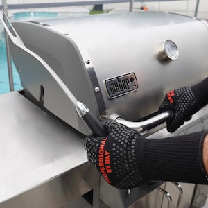 Op de afbeelding: Een persoon met zwarte hittebestendige handschoenen gebruikt een grillgereedschap met een lange steel om de deksel van een zilveren Weber-grill te openen. De grill heeft een ingebouwde thermometer en de tekst "Weber" is zichtbaar aan de voorkant.