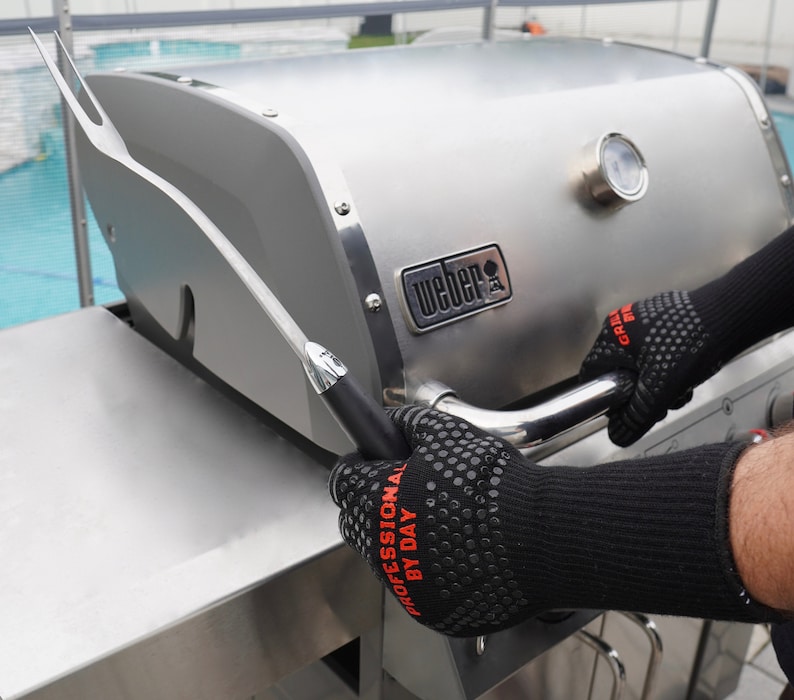 Op de afbeelding: Een persoon met zwarte hittebestendige handschoenen gebruikt een grillgereedschap met een lange steel om de deksel van een zilveren Weber-grill te openen. De grill heeft een ingebouwde thermometer en de tekst "Weber" is zichtbaar aan de voorkant.