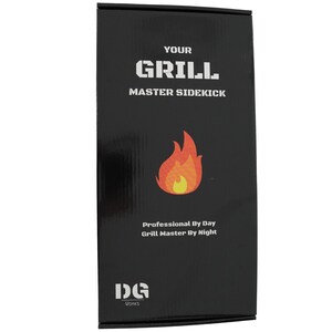 Op de afbeelding: Zwarte doos met witte tekst die "Your Grill Master Sidekick" luidt en een rode en gele vlamgrafiek. De tekst "Professional By Day Grill Master By Night" staat ook op de doos. De onderkant van de doos heeft een wit logo met de tekst "DG Works".