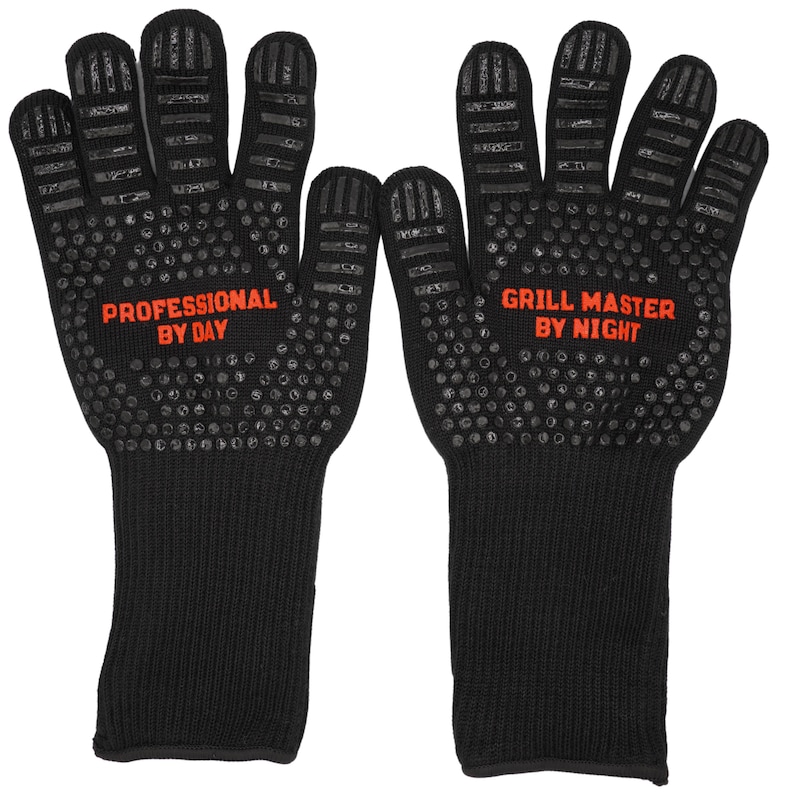 Op de afbeelding: Een paar hittebestendige handschoenen in zwart met rode tekst. De linker handschoen heeft de tekst "Professional by Day" en de rechter handschoen "Grill Master by Night".