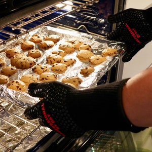 Op de afbeelding: Een bakplaat met vers gebakken chocoladekoekjes wordt uit een oven gehaald met een paar zwarte ovenhandschoenen. De handschoenen hebben de tekst "AVO by PROFESSIONAL" in rood erop gedrukt.