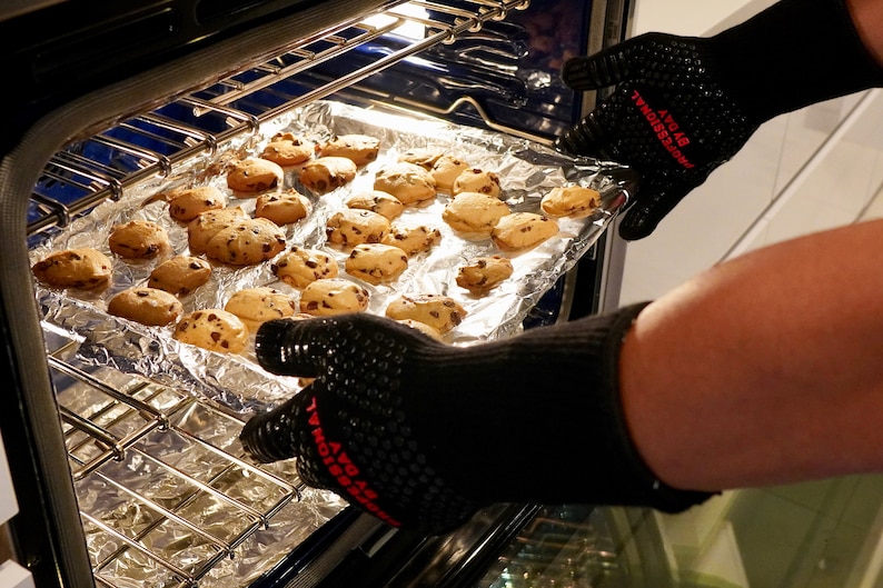 Op de afbeelding: Een bakplaat met vers gebakken chocoladekoekjes wordt uit een oven gehaald met een paar zwarte ovenhandschoenen. De handschoenen hebben de tekst "AVO by PROFESSIONAL" in rood erop gedrukt.