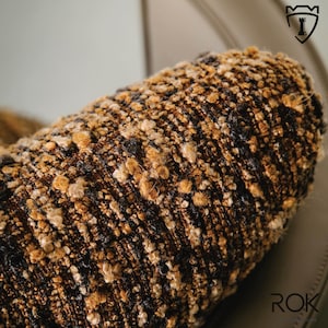 Puede incluir: Primer plano de un coj&iacute;n cil&iacute;ndrico texturizado con una tela tejida marr&oacute;n, negra y beige. La tela tiene una textura nudosa, tipo boucl&eacute;. El coj&iacute;n parece ser parte de un mueble m&aacute;s grande.