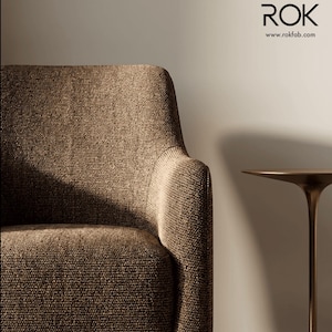 Peut inclure: Gros plan sur un fauteuil marron en tissu texturé. L'accoudoir est incurvé. À droite, une petite table d'appoint dorée avec un plateau rond et un pied fin. Le nom de la marque "ROK" et l'adresse du site web sont visibles.
