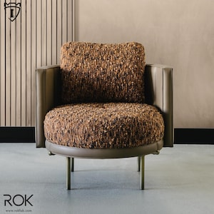 Puede incluir: Sill&oacute;n moderno con asiento y respaldo redondeados, tapizado en tela marr&oacute;n texturizada. La silla presenta un perfil bajo, con ribetes y reposabrazos de cuero gris. Las patas son delgadas, met&aacute;licas y rectas.