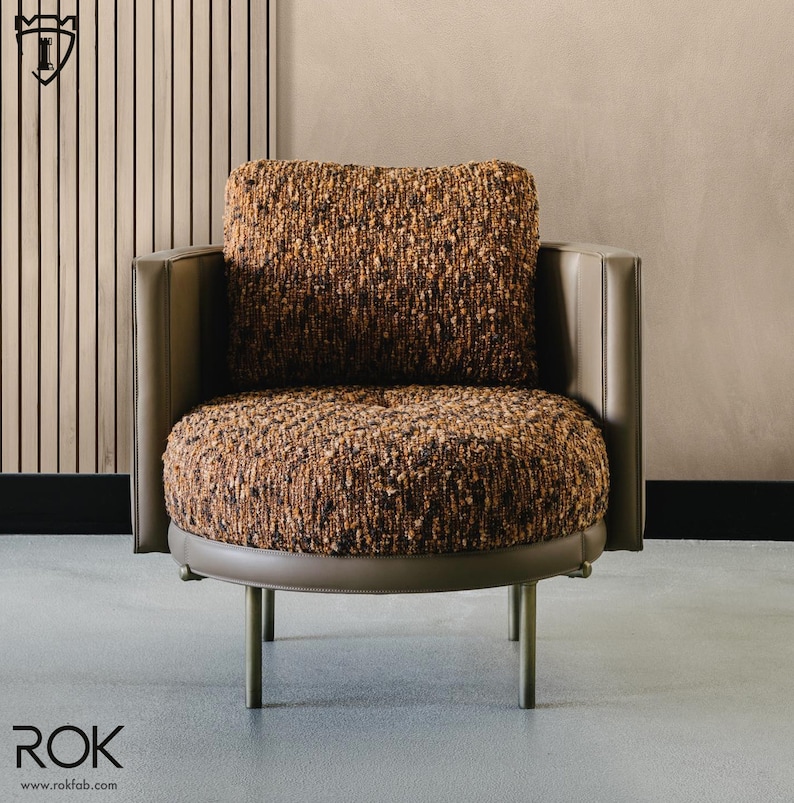 Puede incluir: Sill&oacute;n moderno con asiento y respaldo redondeados, tapizado en tela marr&oacute;n texturizada. La silla presenta un perfil bajo, con ribetes y reposabrazos de cuero gris. Las patas son delgadas, met&aacute;licas y rectas.