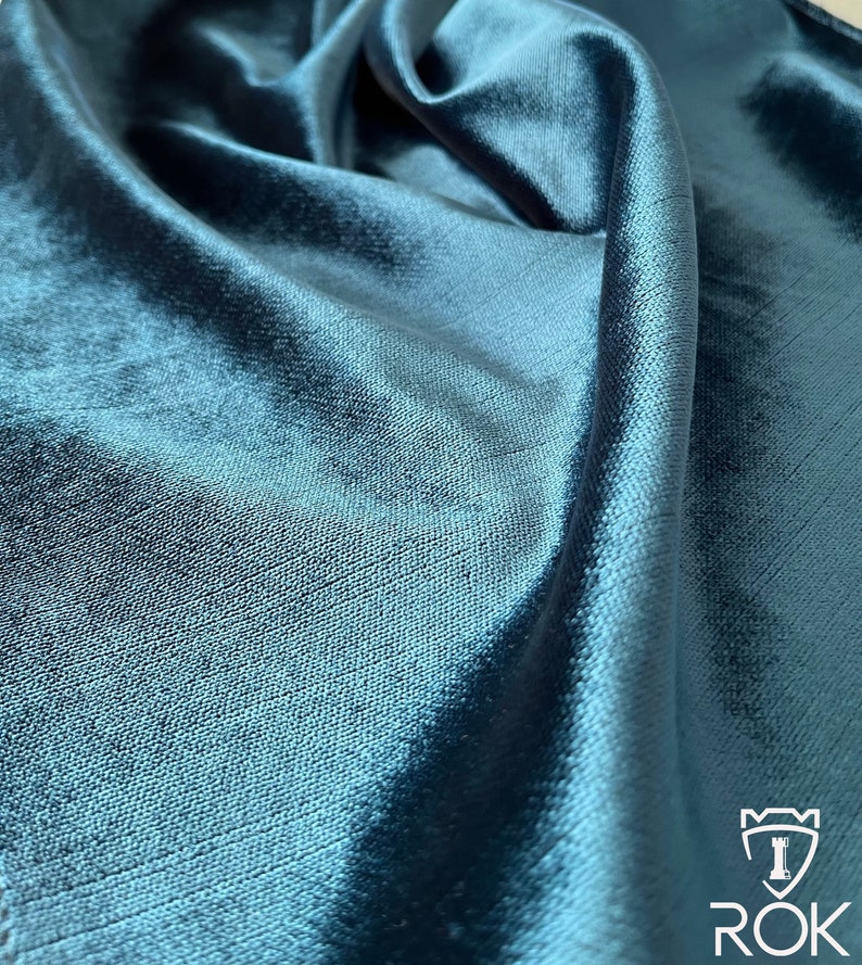 May include: Close-up of a teal blue fabric with a smooth, silky texture. The fabric is folded and draped, showcasing its luxurious feel.