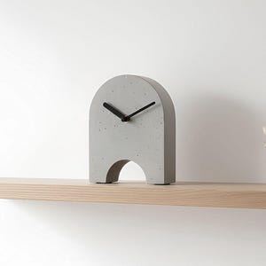 May include: A modern, gray concrete clock with black hands, set on a light wood shelf. The clock has an arched top and a cutout at the base. A small glass vase with dried flowers is to the right.