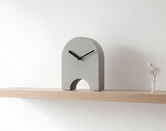 May include: A modern, gray concrete clock with black hands, set on a light wood shelf. The clock has an arched top and a cutout at the base. A small glass vase with dried flowers is to the right.