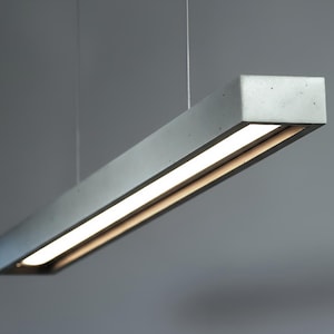 May include: A rectangular, gray concrete pendant light fixture with a bright, linear LED light source. The modern design features a minimalist aesthetic, suspended from the ceiling by thin wires. The light fixture is suitable for interior lighting.