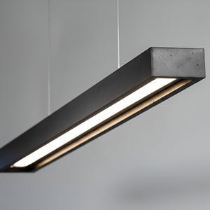 May include: A modern, rectangular pendant light fixture with a dark gray, concrete-like finish. The light has a bright, white LED strip running along the bottom. The fixture is suspended from the ceiling by thin wires.