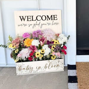 Puede incluir: Un letrero de madera con el texto "WELCOME we're so glad you're here" y un arreglo floral en una maceta con el texto "baby in bloom".