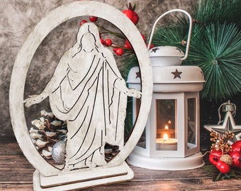 May include: White wooden cutout of a religious figure standing with arms outstretched, set against a rustic background with a white lantern and red berries.