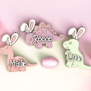May include: Three pastel-coloured dinosaur ornaments with bunny ears and personalised names: Mallie, Reece, and Linc. The ornaments are surrounded by pastel-coloured Easter eggs.