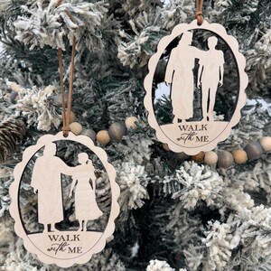 Puede incluir: Dos adornos navideños de madera con borde festoneado, cada uno con un diseño recortado de dos figuras y las palabras "WALK with ME". Los adornos cuelgan de cordones marrones sobre un fondo de árbol de Navidad nevado.