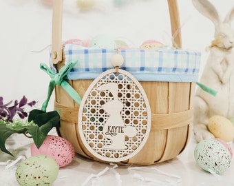May include: A wooden Easter basket with a blue and white checkered liner, filled with pastel colored eggs. A wooden egg-shaped ornament with a bunny cutout and the name "Kayte" is hanging from the basket.