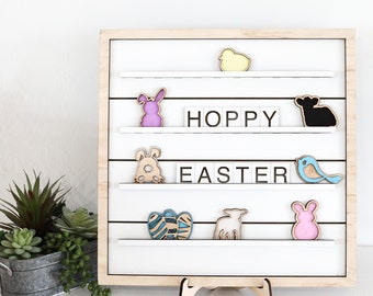 May include: A wooden sign with a white background and a wooden frame. The sign has a series of shelves with various Easter-themed decorations, including a chick, a bunny, a lamb, an egg, and a bird. The sign also has letter tiles spelling out "Hoppy Easter".