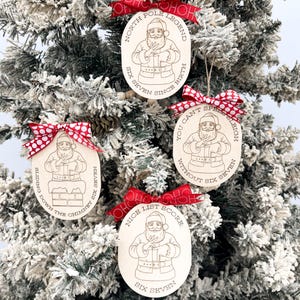 Könnte beinhalten: Vier hölzerne Weihnachtsschmuckstücke mit rot-weiß gepunkteten Schleifen hängen an einem schneebedeckten Baum. Jedes Ornament zeigt eine Weihnachtsmann-Illustration und den Text: "North Pole Legend", "Sliding Down the Chimney", "You Can't Spell Sleigh" und "Nice List Score".