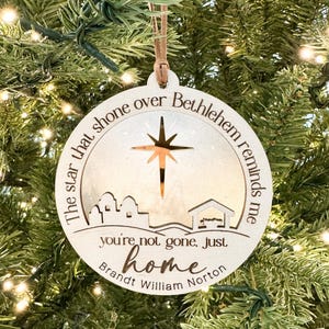 以下が含まれることがあります： 星とベツレヘムのシーンが切り抜かれた、丸い木製のクリスマスオーナメント。オーナメントには「ベツレヘムの上に輝いた星は私に思い出させてくれる、あなたは去ったのではなく、ただ家にいるだけだ」というテキストと「Brandt William Norton」という名前が書かれています。