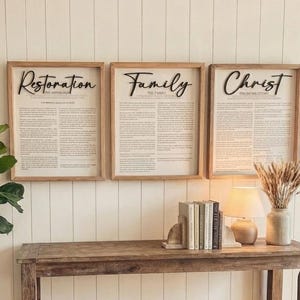 Puede incluir: Tres obras de arte enmarcadas con las palabras "Restoration", "Family" y "Christ" en escritura negra. Los marcos son de color marrón claro y el fondo es de color crema. Debajo del arte hay una consola de madera con libros, una lámpara y un jarrón.