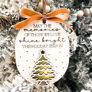 May include: White wooden Christmas ornament with a gold glitter tree and the text "May the memories of those we love shine bright this holiday season".