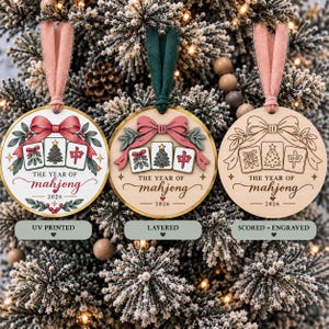 Peut inclure: Trois décorations de Noël rondes en bois avec un motif de tuiles de mahjong, un nœud, du gui et le texte THE YEAR OF mahjong 2026. Les ornements sont présentés en styles imprimé UV, superposé, et gravé avec des rubans en tissu.