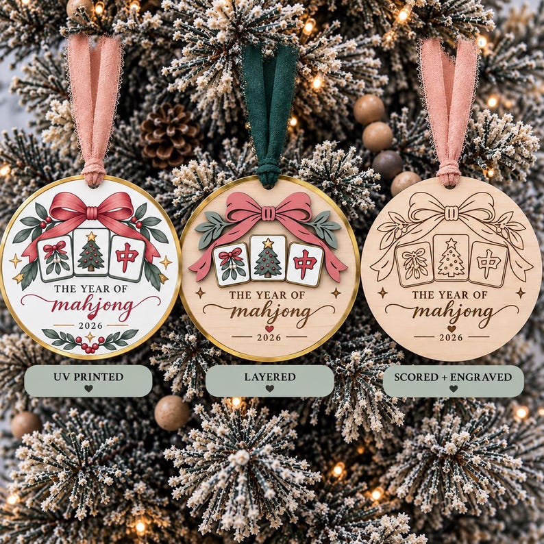 May include: Three round wooden Christmas ornaments featuring a mahjong tile design with a bow, mistletoe, and the text THE YEAR OF mahjong 2026. The ornaments are displayed in UV printed, layered, and scored and engraved styles with fabric ribbons.