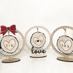 May include: Three wooden ornaments with a heart-shaped design and the initials "T & K" and "T ♥ K". The ornaments are hanging from a rope and are on a wooden stand. The middle ornament has the word "love" written on the stand.