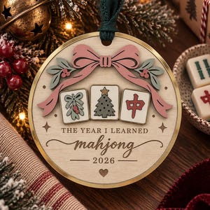 May include: A round wooden Christmas ornament featuring a pink bow, three mahjong tiles with mistletoe, a Christmas tree, and a red character, with the text THE YEAR I LEARNED mahjong 2026, suspended by a dark green ribbon.