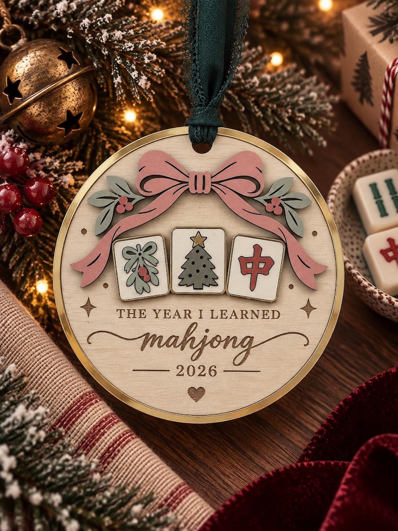 May include: A round wooden Christmas ornament featuring a pink bow, three mahjong tiles with mistletoe, a Christmas tree, and a red character, with the text THE YEAR I LEARNED mahjong 2026, suspended by a dark green ribbon.