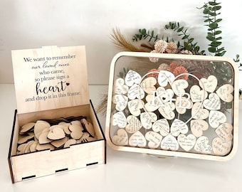 May include: A wooden box with a sign that reads "We want to remember our loved ones who came, so please sign a heart and drop it in this frame." The box is filled with small wooden hearts. Next to the box is a clear glass frame with a wooden heart-shaped frame inside. The frame is filled with wooden hearts that have been signed by guests. The hearts are arranged in a heart shape. The frame is on a wooden stand.