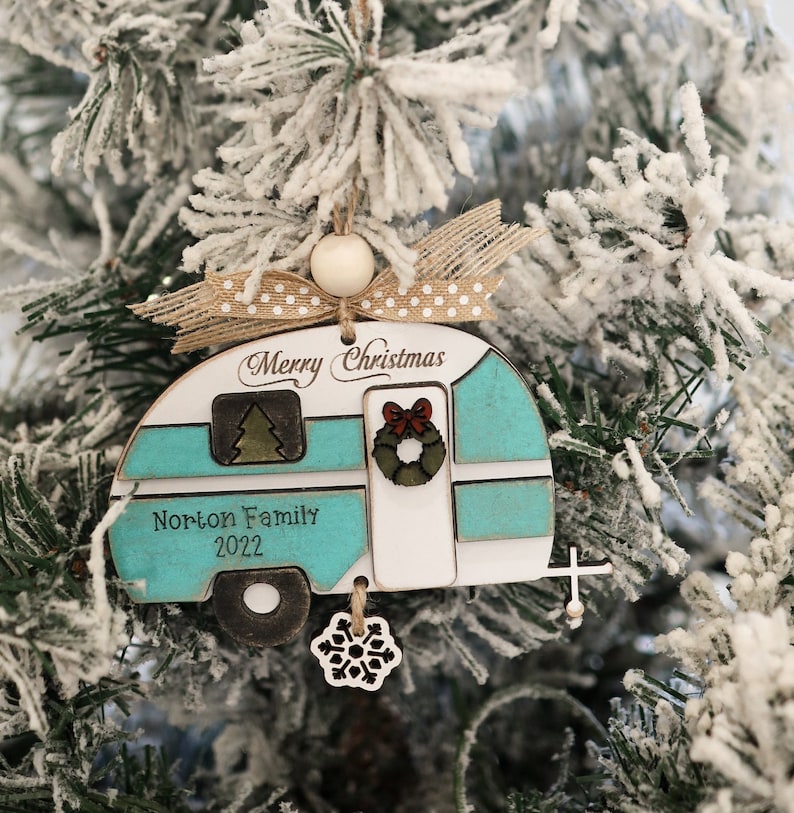 May include: A teal and white wooden Christmas ornament shaped like a camper with the text "Merry Christmas" and "Norton Family 2022". The ornament has a brown ribbon with white polka dots and a white snowflake charm.