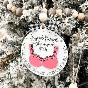 May include: A white round ornament with a pink bra cutout and the text "A good friend is like a good bra + supportive, never leaves you hanging, & always close to your heart +"