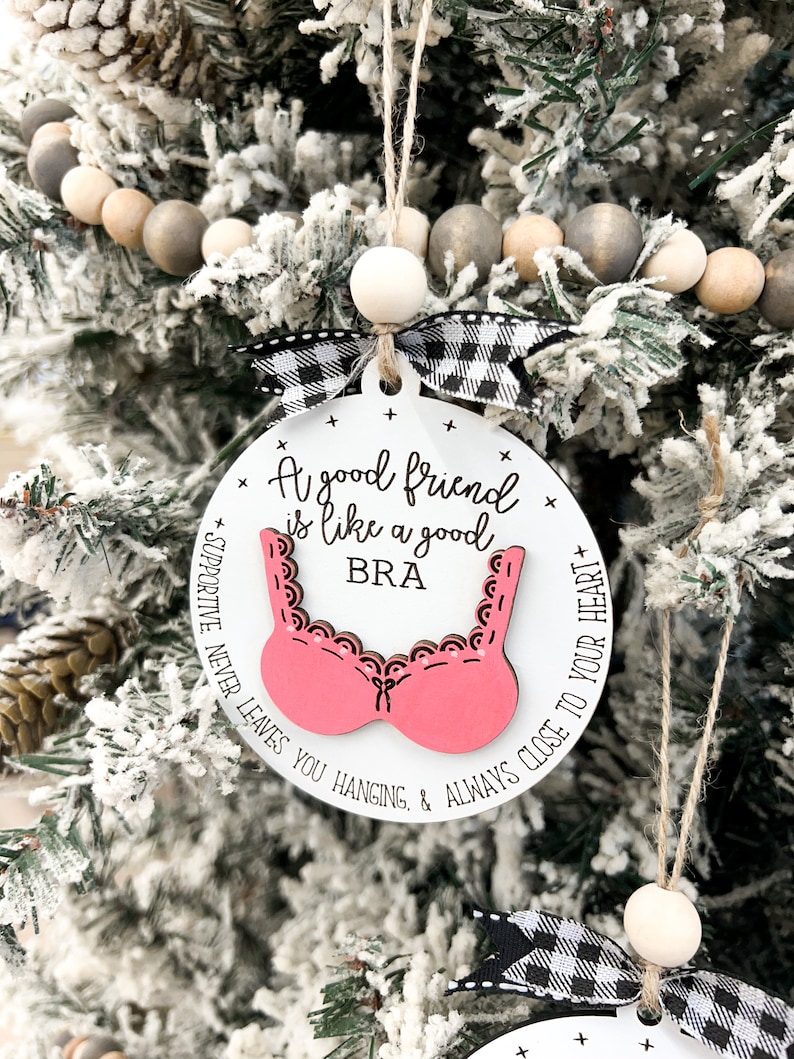 May include: A white round ornament with a pink bra cutout and the text "A good friend is like a good bra + supportive, never leaves you hanging, & always close to your heart +"